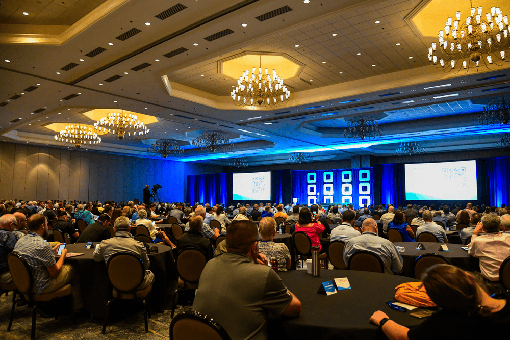 Bright conference hall packed with professionals, Chris Hood AI Keynote Speaker, attending a corporate event, large screens displaying presentation content, elegant chandeliers overhead, showcasing a professional business conference setting.