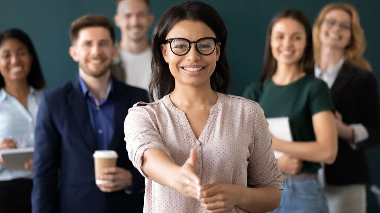Bright smiling woman in glasses extending hand for a handshake with diverse business team in background.