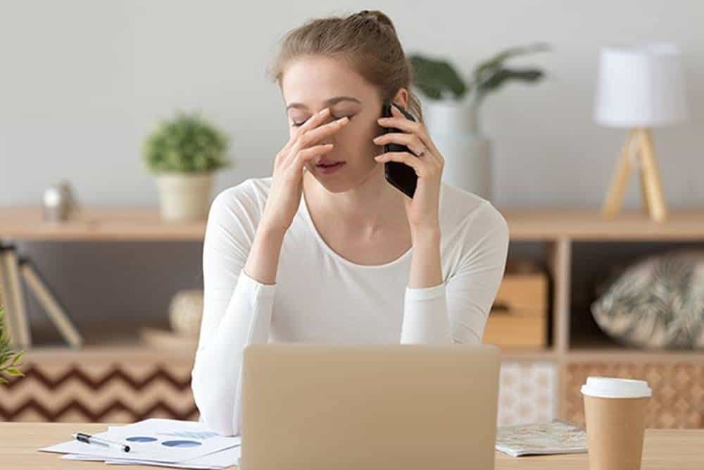 Overwhelmed woman talking on phone, holding forehead, working from home, stressed, in a modern home office.