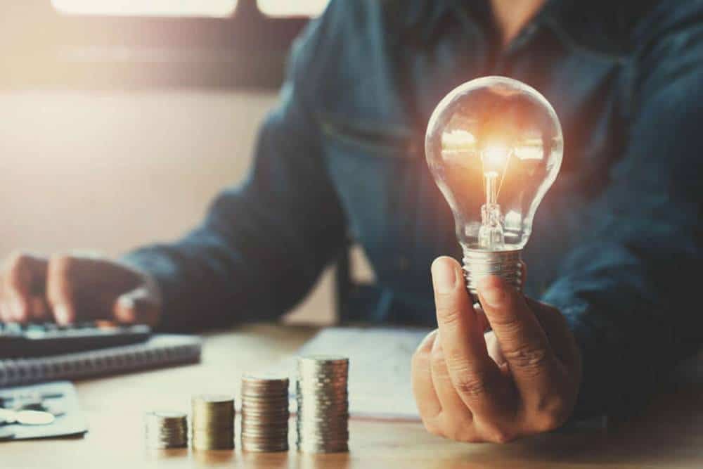 Bright idea concept with a person holding a glowing light bulb, stacks of coins, and a calculator representing innovation, finance, and entrepreneurship.