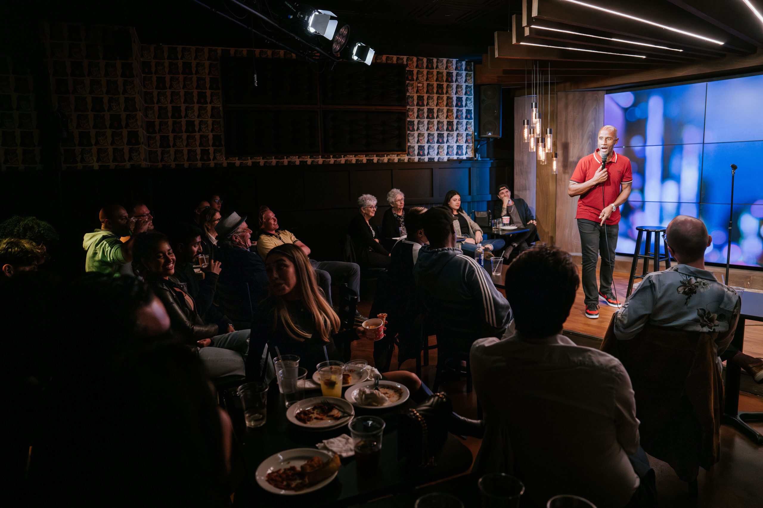 Comedian in red shirt on stage at The Comedy Bar with audience at tables