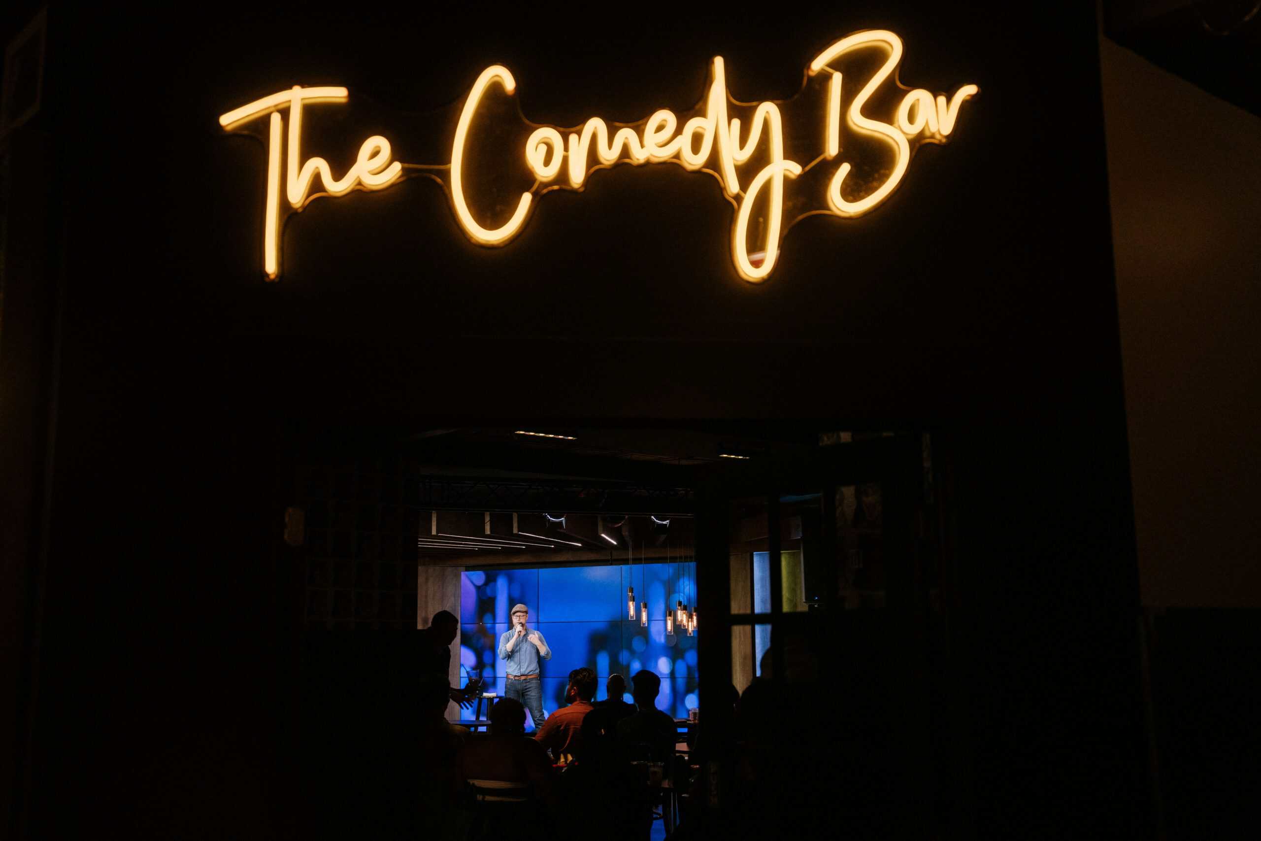 Neon Comedy Bar sign with comedian performing on stage below