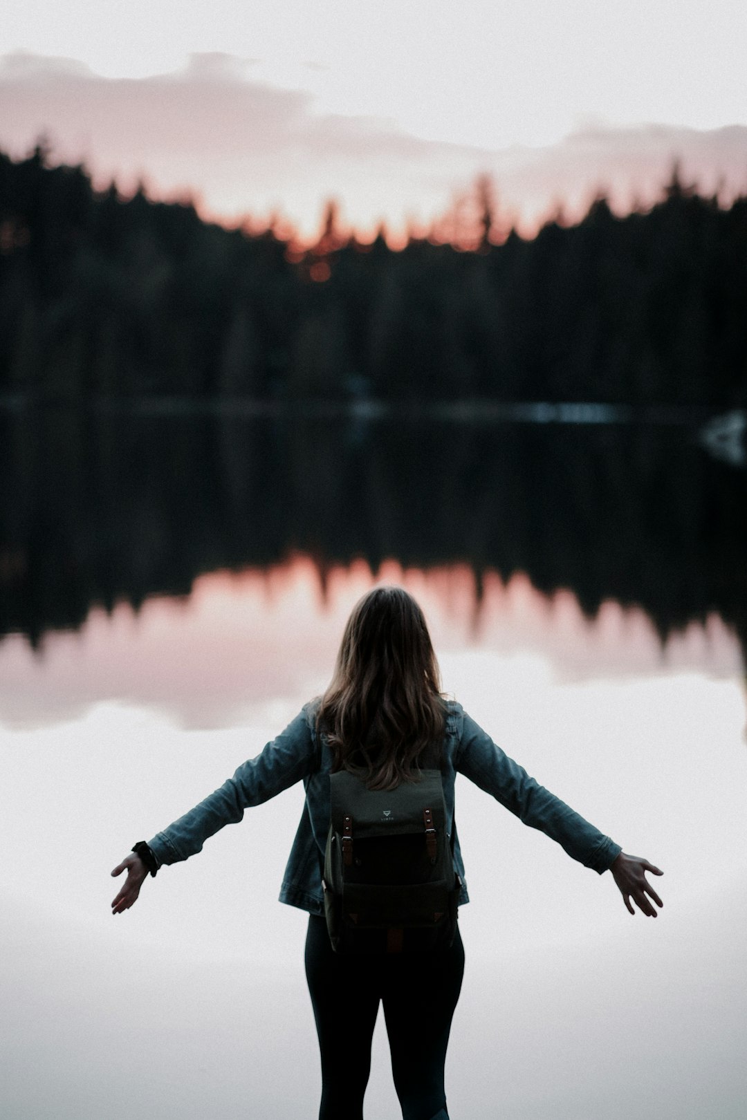Woman standing with arms open wide in front of lake looking calm and refreshed.