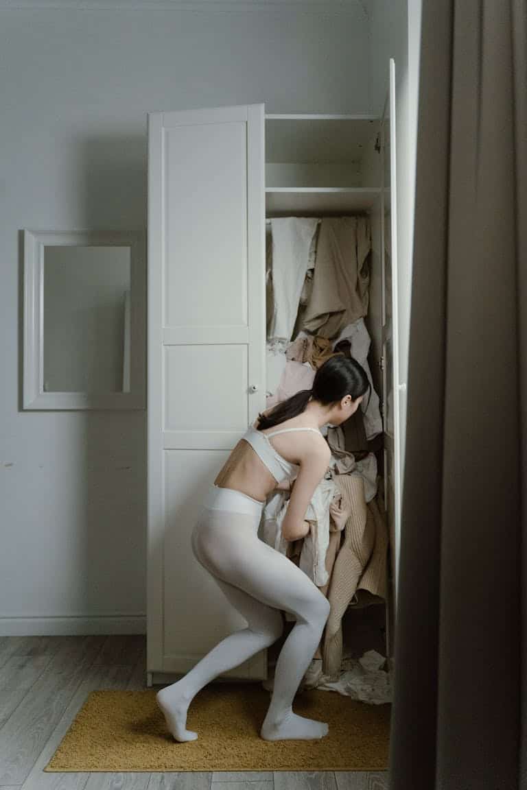 A woman in white leggings organizes a cluttered wardrobe in a minimalist room.