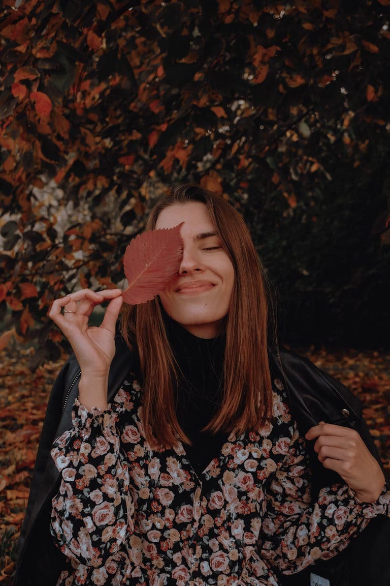 Woman in floral dress smiling with leaf over eye in autumn setting.