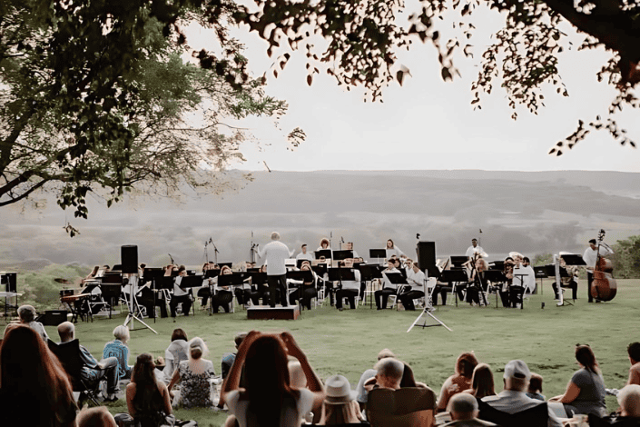 DCA Saxophone Orchestra performing outdoors on a lush green lawn with scenic hills in the background, under a tree canopy, during a live concert with an audience enjoying the music in the evening.