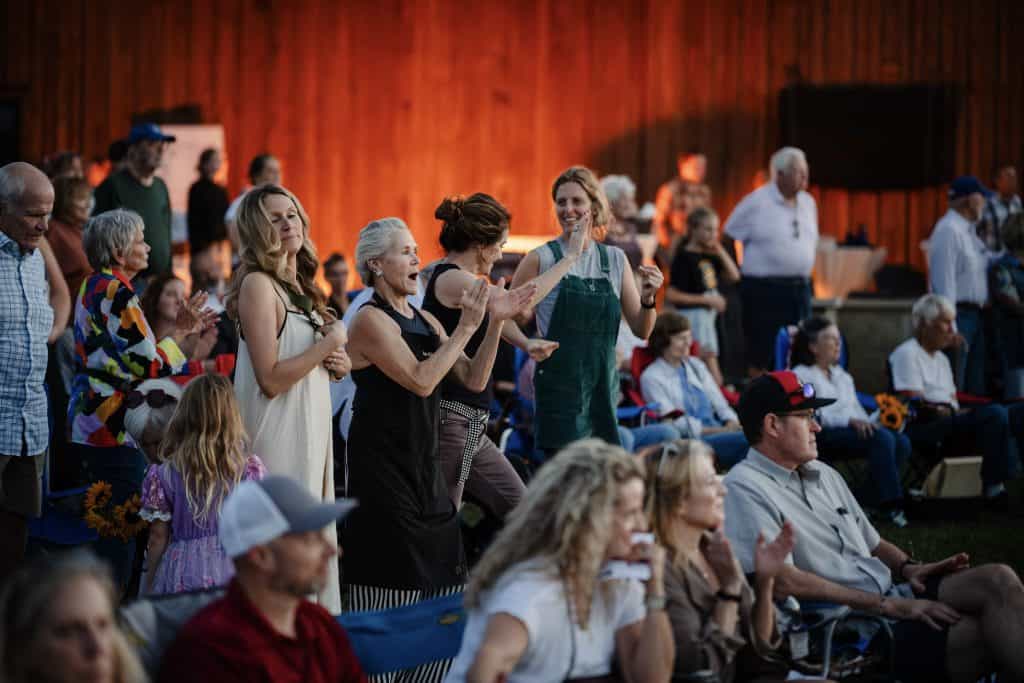People of various ages and backgrounds are engaged in a lively outdoor event, some standing and clapping, others seated, with a warm wooden backdrop and evening lighting.