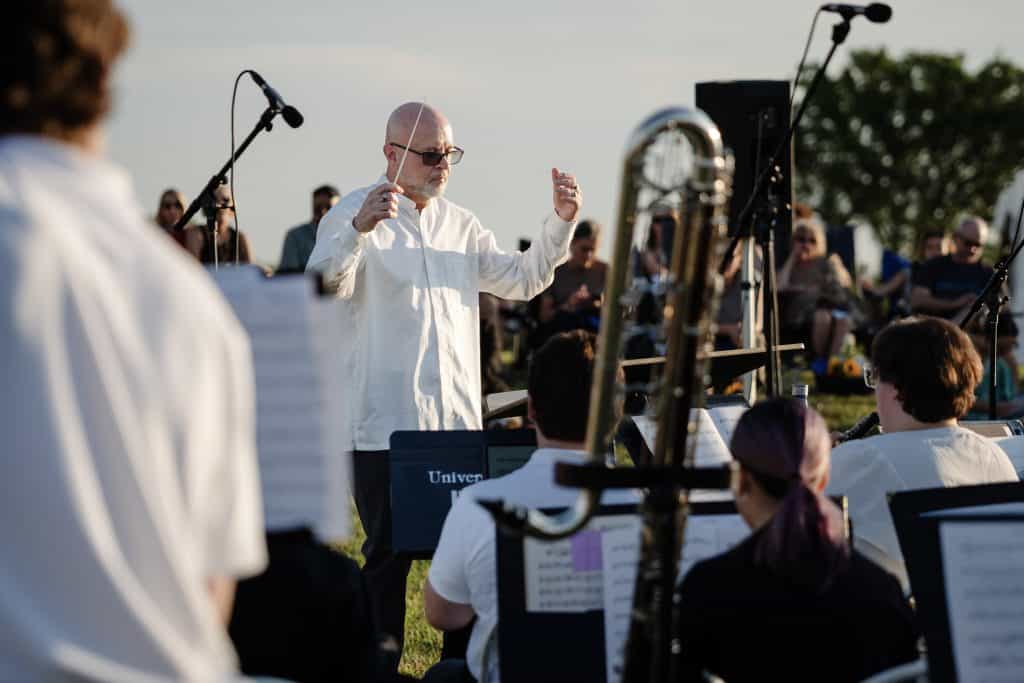 Conductor directing an outdoor orchestra during a sunset concert with musicians and audience in the background.