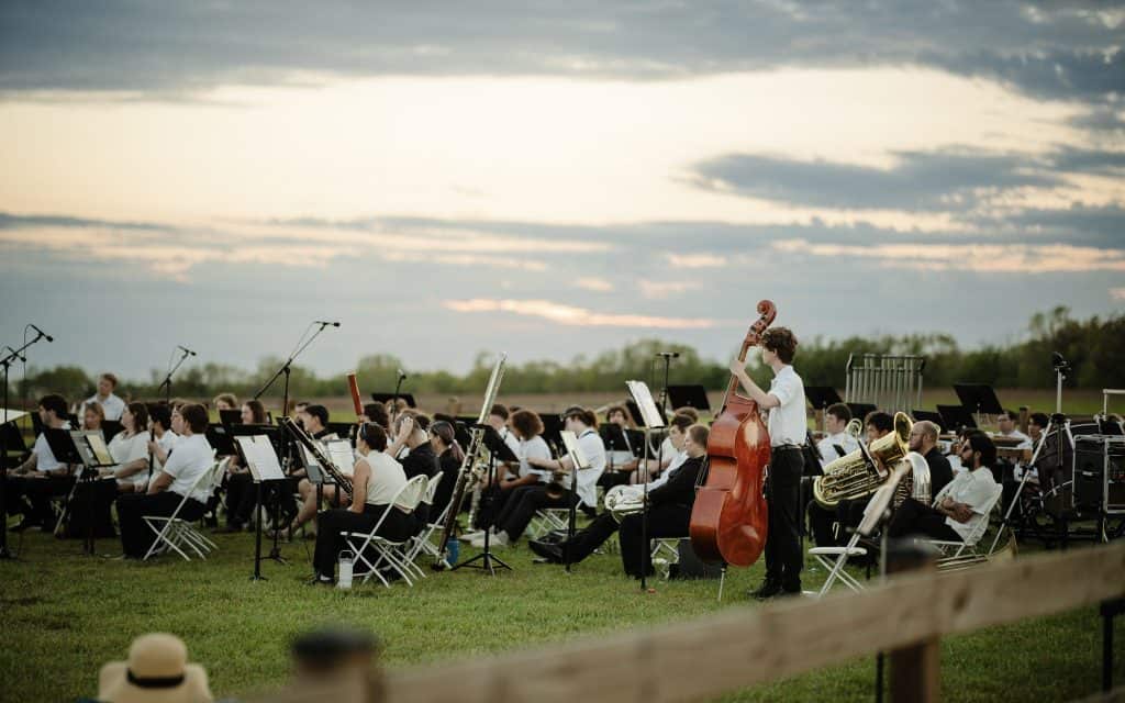 A large outdoor orchestra performing on a grassy field during sunset, with musicians playing various instruments including a double bass, under a partly cloudy sky.