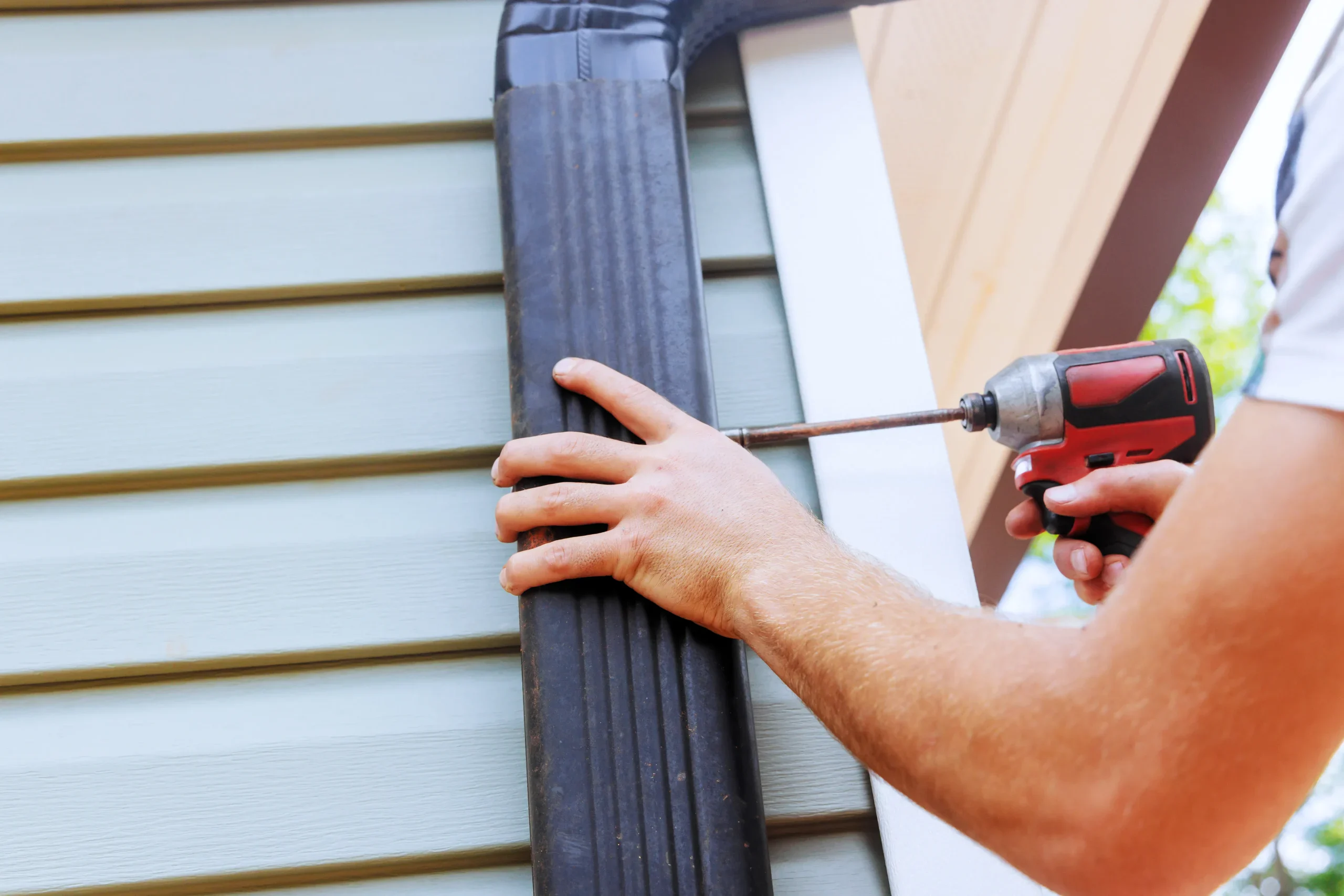 Man installing a black downspout on a house with a power drill