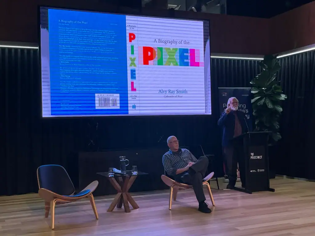 Dr. Alvy Ray Smith standing behind a podium speaking at The Precinct stadium, with the projection screen behind him displaying his book cover for A Biography of the Pixel.