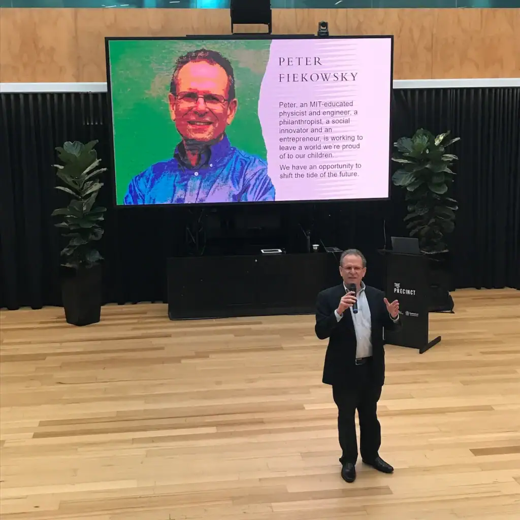 Peter Fiekowsky standing on stage presenting with an active hand gesture, in front of a large projection screen displaying his headshot and professional background profile.