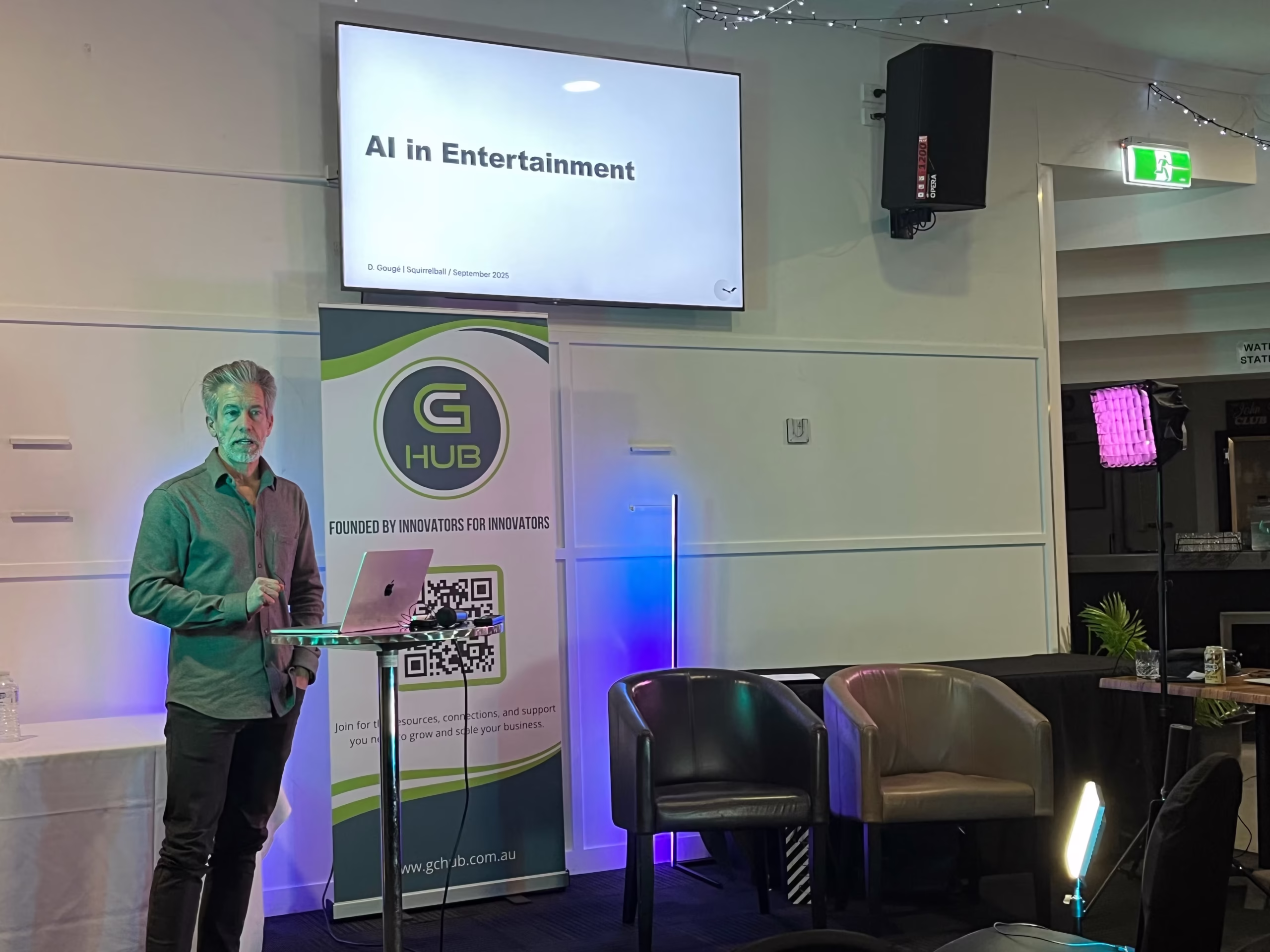 Dave Gougé standing behind a high cocktail table with his laptop, delivering an executive briefing in front of an AI in Entertainment screen display and a Gold Coast Innovation Hub banner.