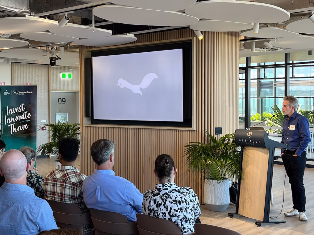 Wide right-angle view of Dave Gougé standing behind the podium listening to an audience question, with a prominent Sunshine Coast Council banner displaying the text Invest, Innovate, Thrive visible on the far left.