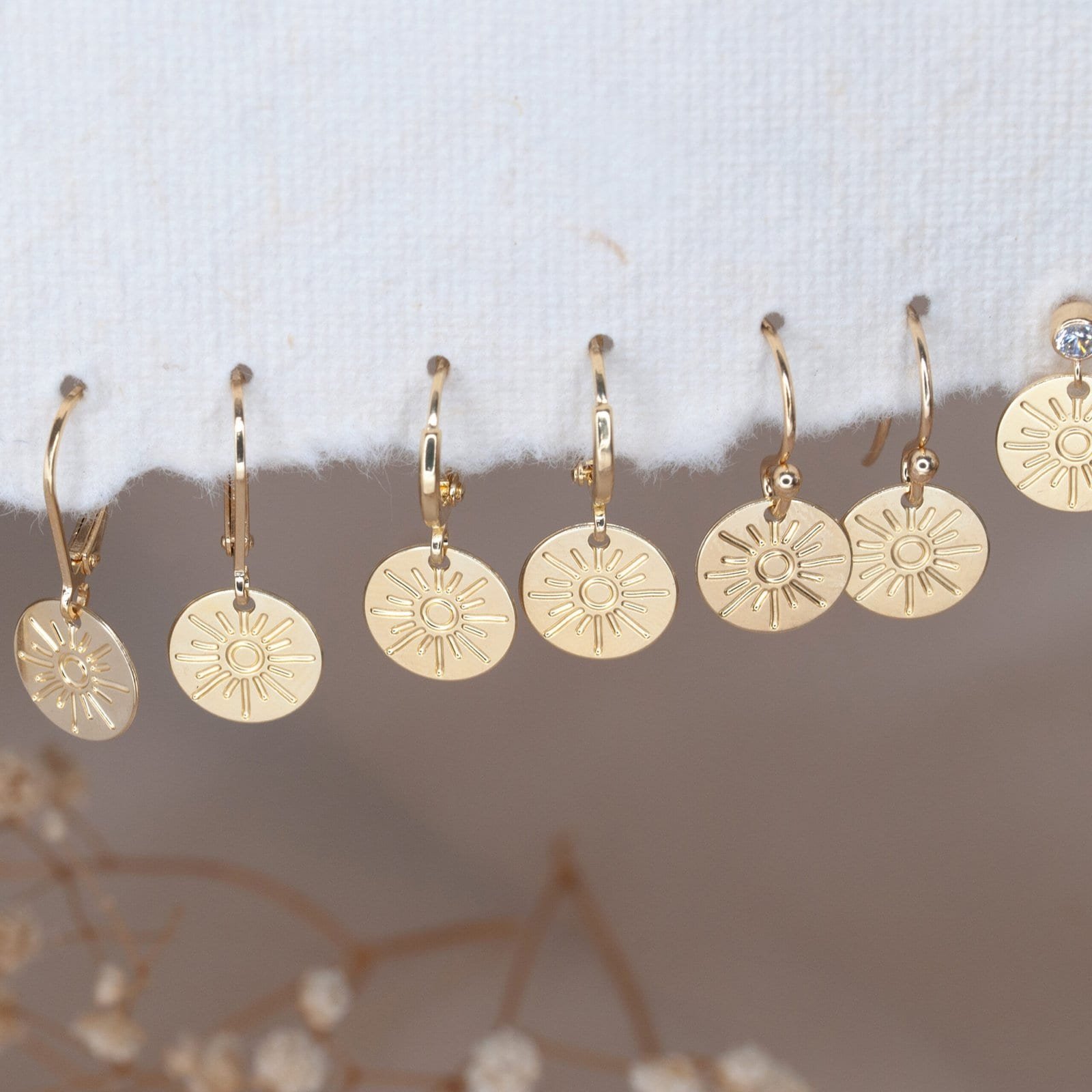 Various gold sun disc earring styles, including hoops, hooks and leverbacks, arranged on soft fabric