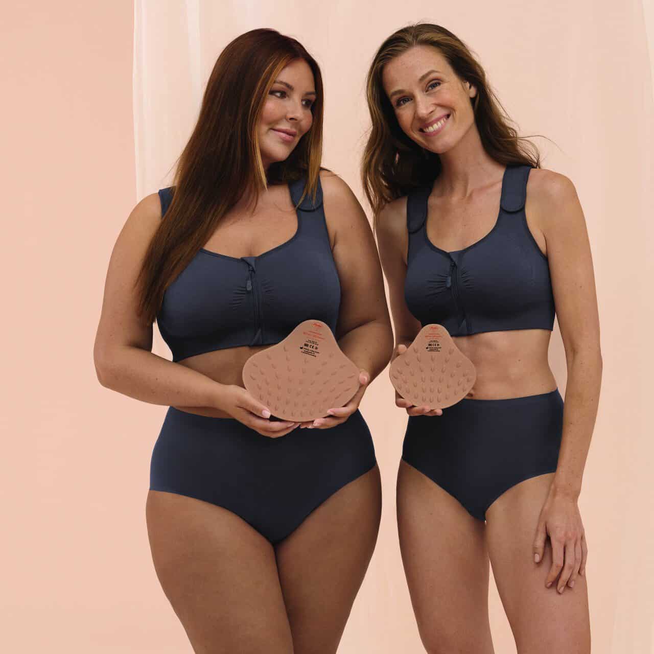 Two women wearing matching dark blue post-surgery bras, each holding a peach silicone breast prosthesis smiling at the camera, demonstrating mastectomy bras.",