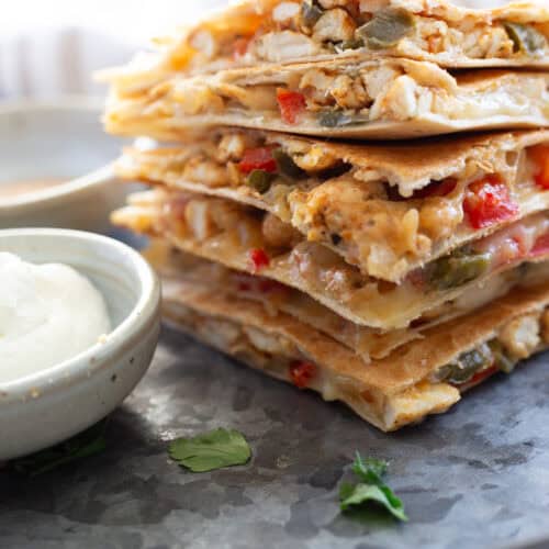 Six chicken quesadillas stacked near a small bowl of sour cream for dipping.