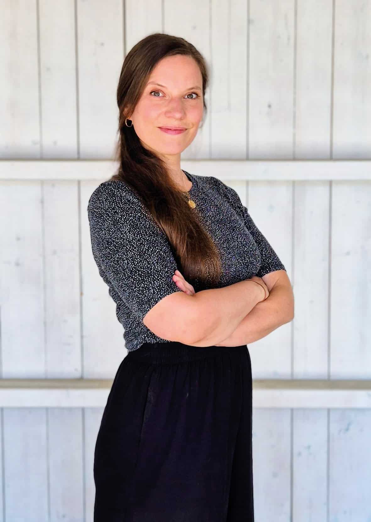 Christiane steht mit verschränkten Armen freundlich lächelnd nach rechts geneigt vor einer weißen Holzwand. Sie hat langes braunes Haar und trägt ein schwarz weiß gepunktetes Shirt.