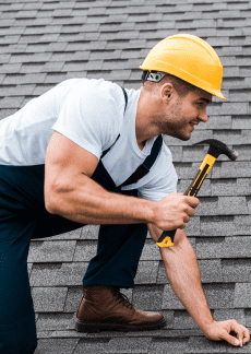 Man in hard hat and overalls hammering a nail on a shingled roof