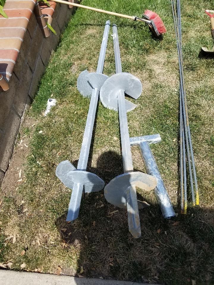 Four metal ground screws and rebar on grass next to a brick wall.