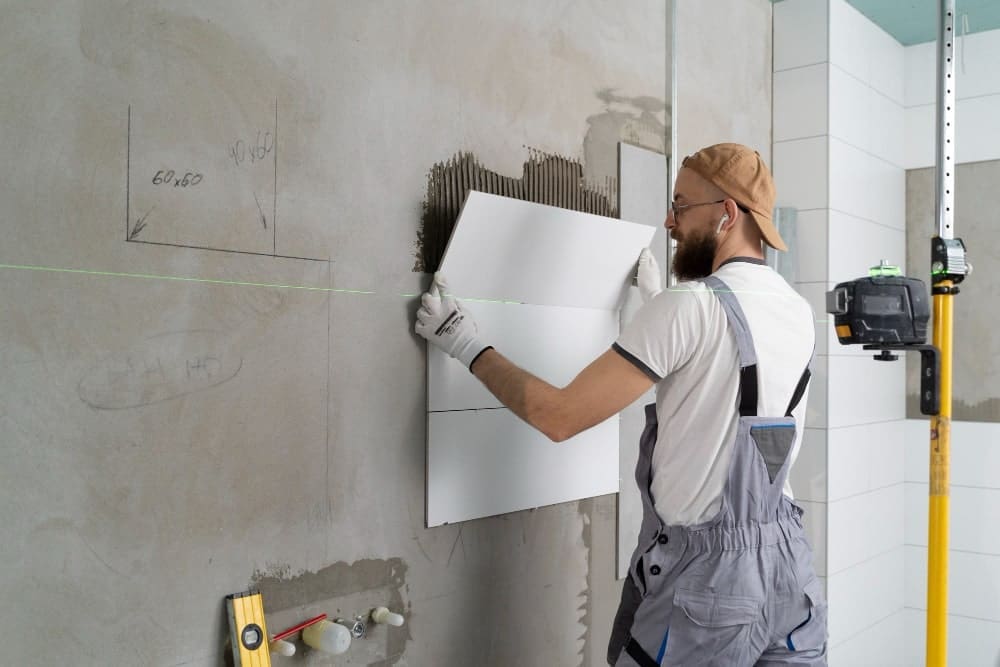 Man installing white tiles on a wall with adhesive and a laser level