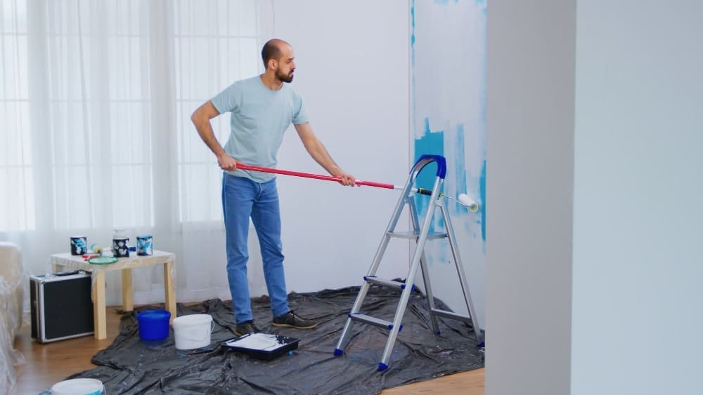 Man painting a wall blue with a roller, standing on a drop cloth.