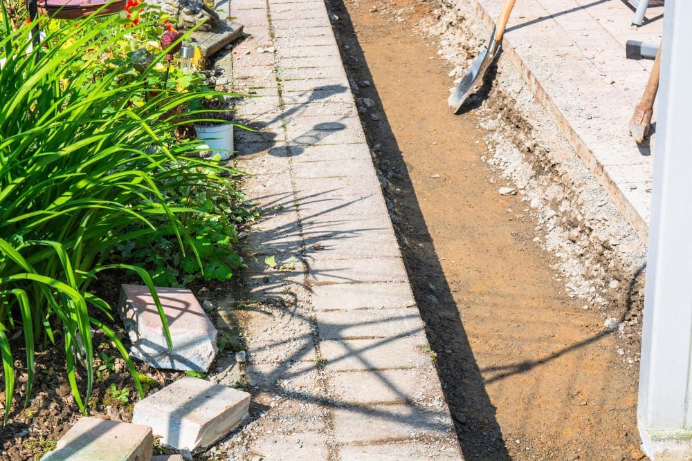 A narrow trench dug alongside a brick pathway and green plants.