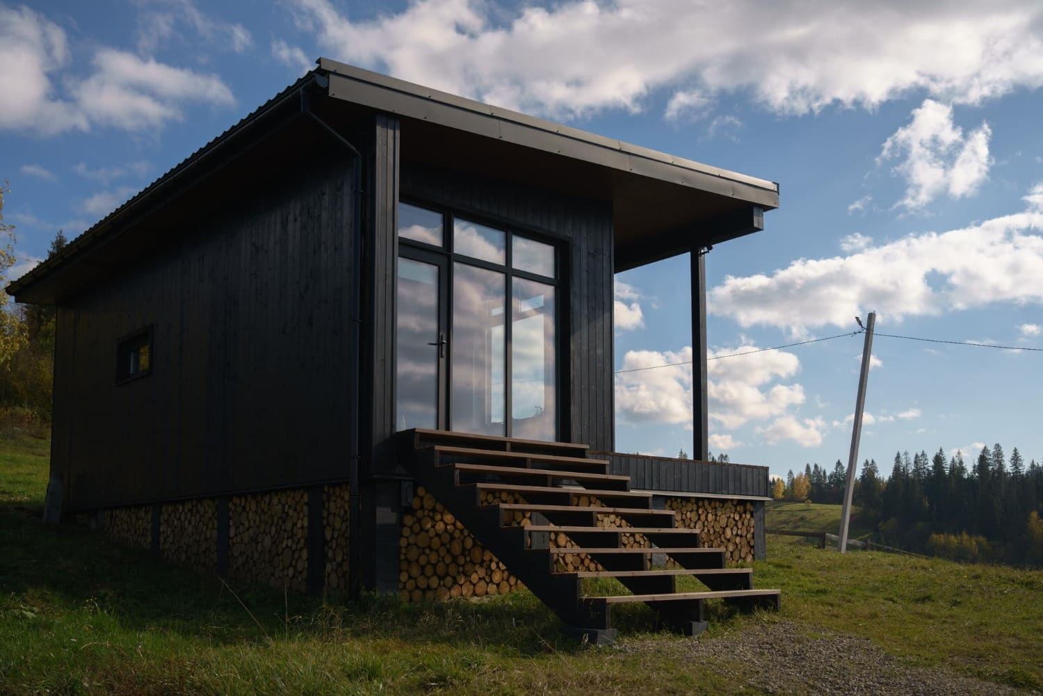 Modern black cabin with wood pile base and wooden steps under a cloudy sky.