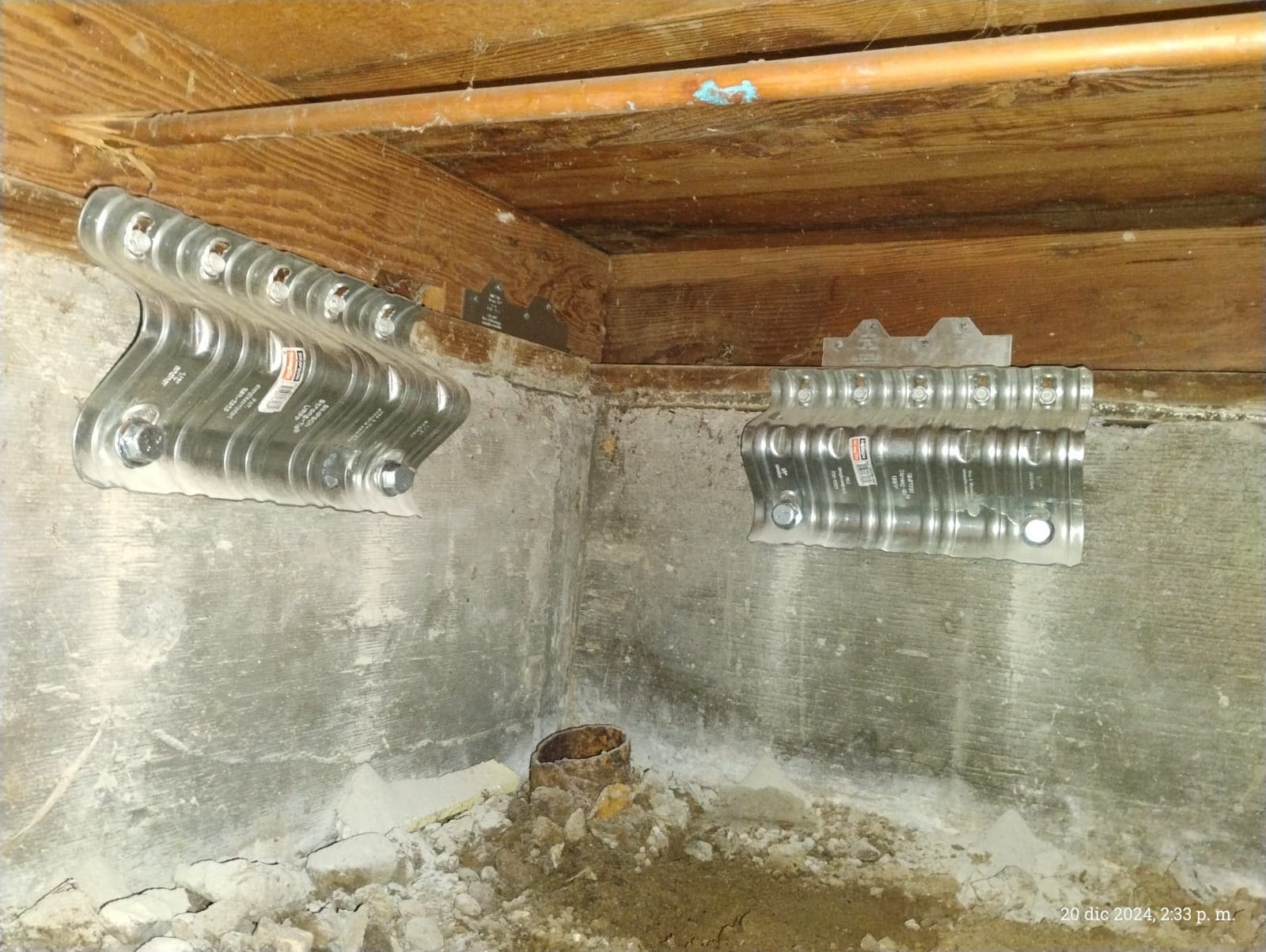 Metal seismic retrofitting plates bolted to a concrete foundation wall and wooden beams.