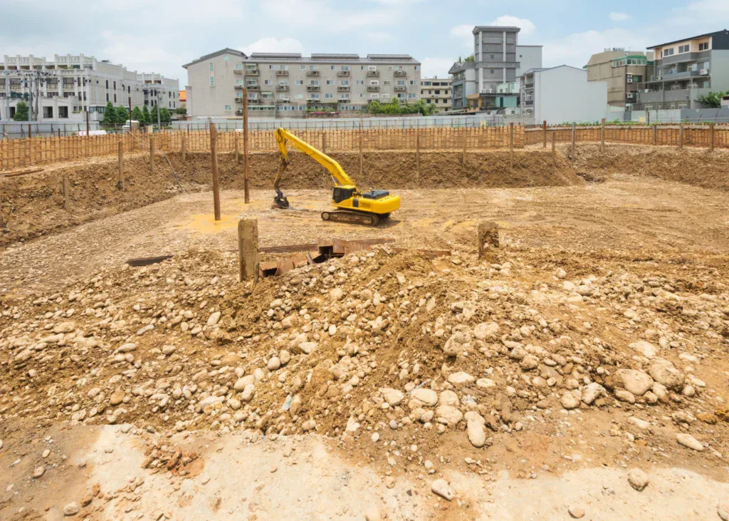 Yellow excavator digging in a large construction pit with buildings in background