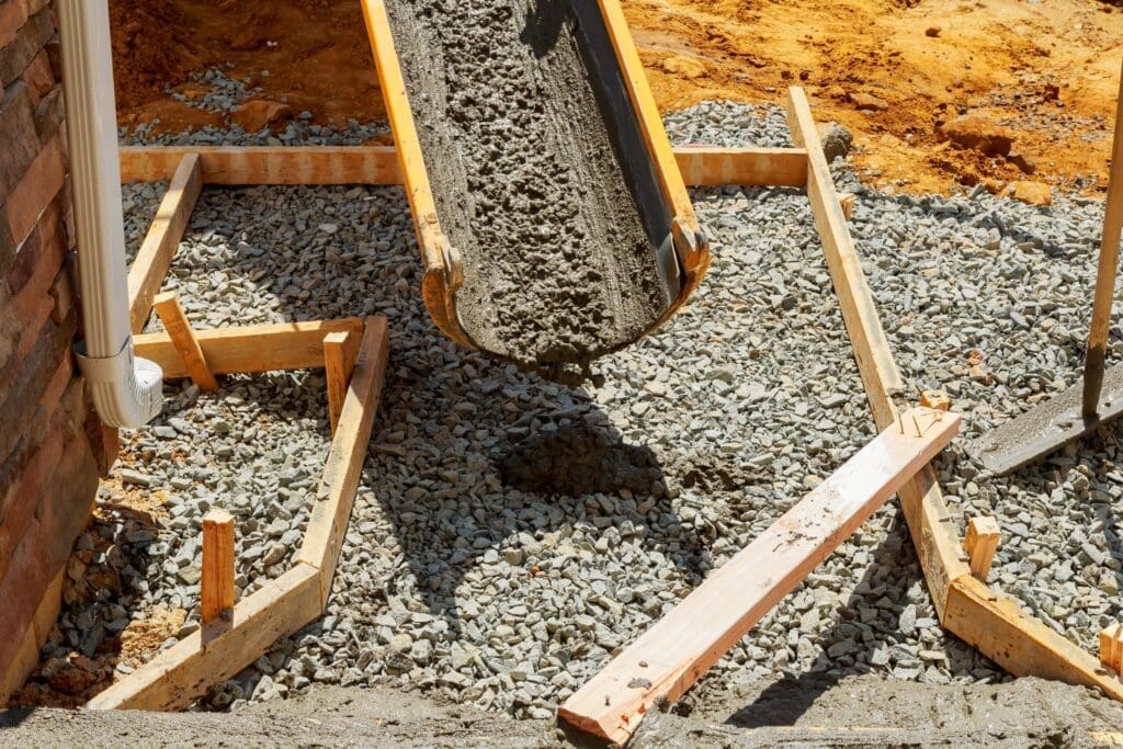 Concrete being poured from a chute into a wooden frame on a gravel base.