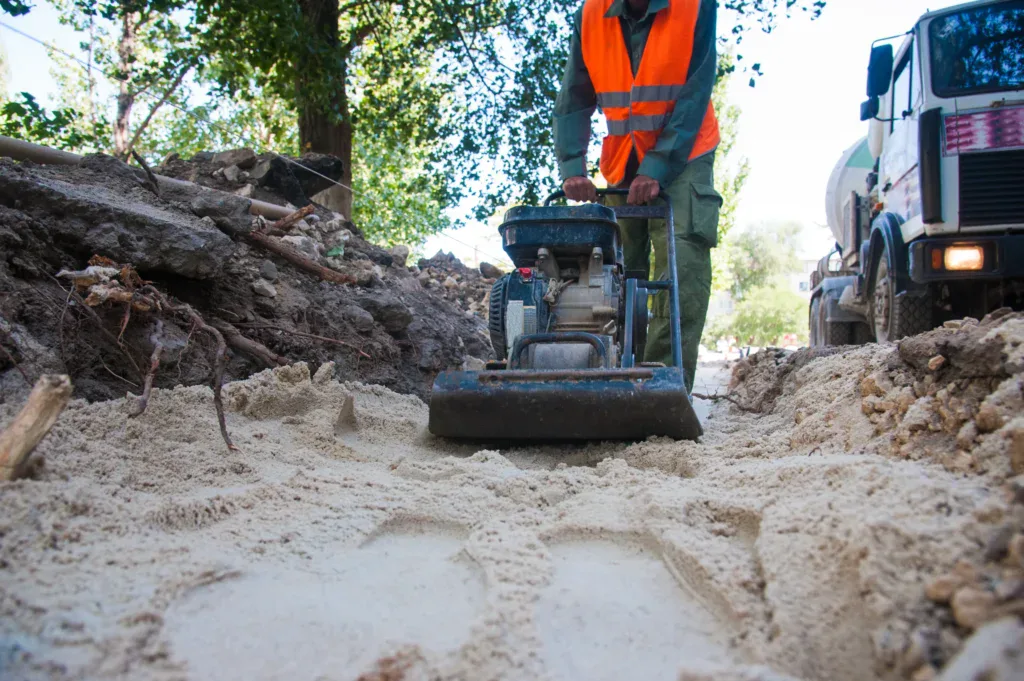 Worker in orange vest compacting sand with a vibratory plate compactor.