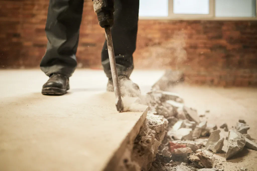 Person using a jackhammer to break concrete, creating dust and debris.