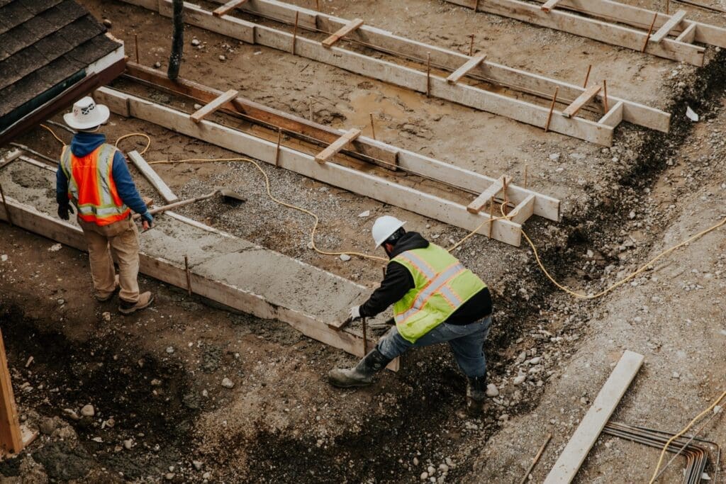 Two construction workers pouring concrete into wooden forms on a building site.