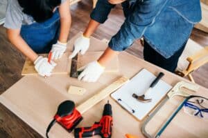 High-angle view of two individuals working together on carpentry; one marks wood using a square while the other assists, surrounded by tools.