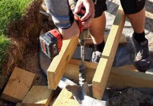 Close-up of hands operating an impact wrench to drive a screw pile. He is standing in between thick wooden beams, so the screw piles can be attached to the structure. He is working on an outdoor home improvement project. How tp prevent deck heave and settlement.