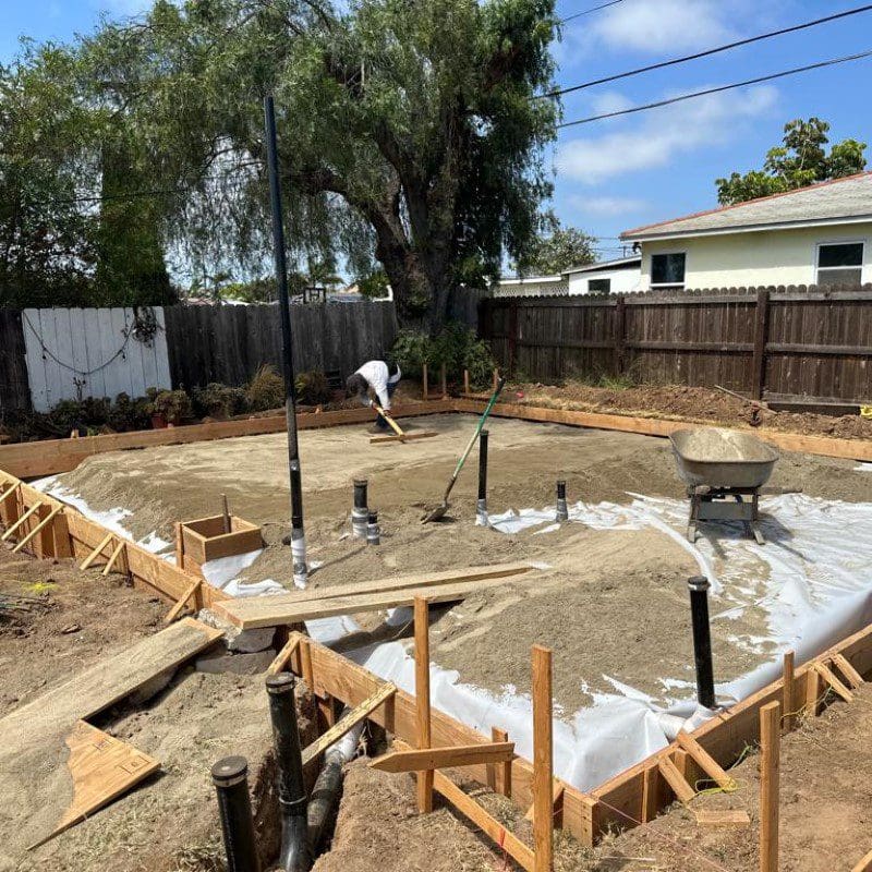 Construction site with wooden forms, sand, pipes, and a worker leveling sand.