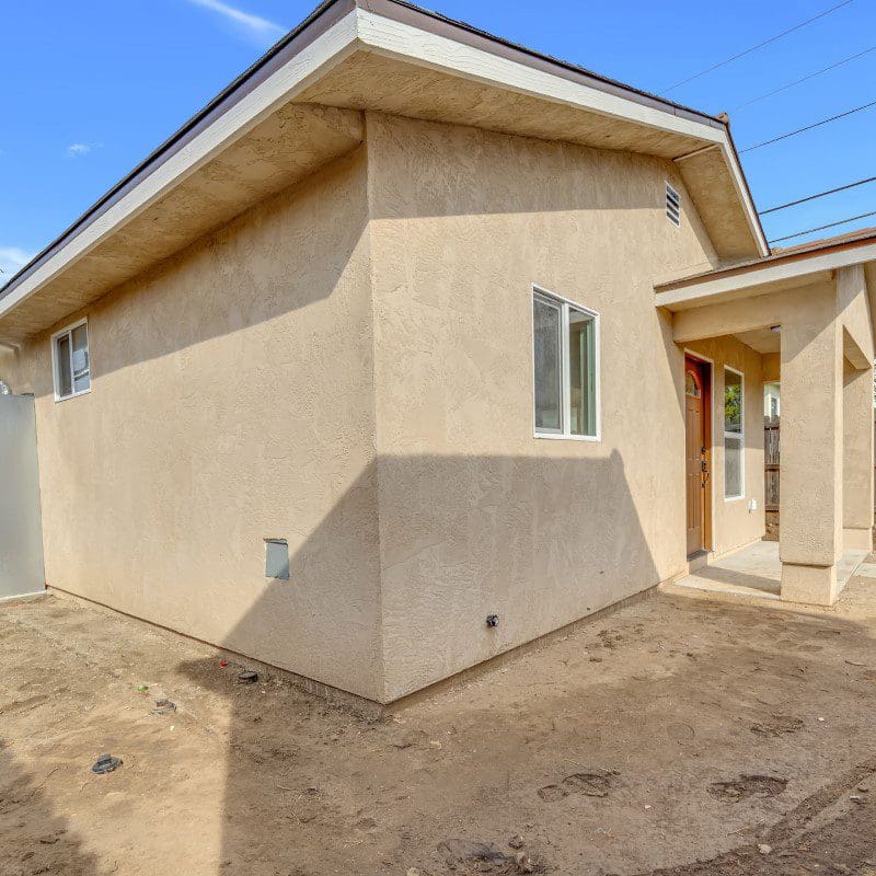 Exterior of a small, tan stucco house with a brown roof and dirt yard.