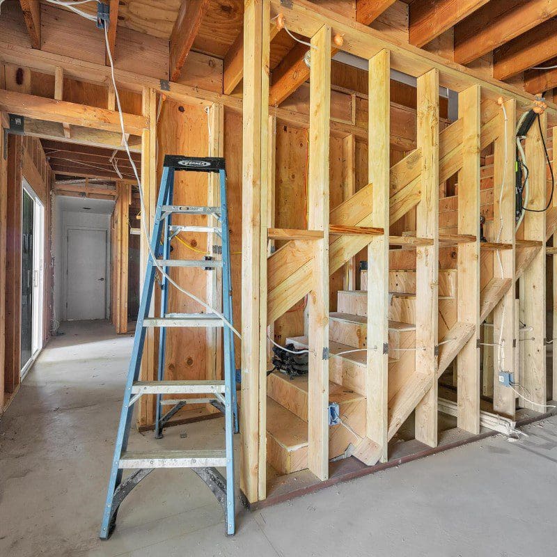 Interior view of a house under construction with exposed wooden framing and a blue ladder.