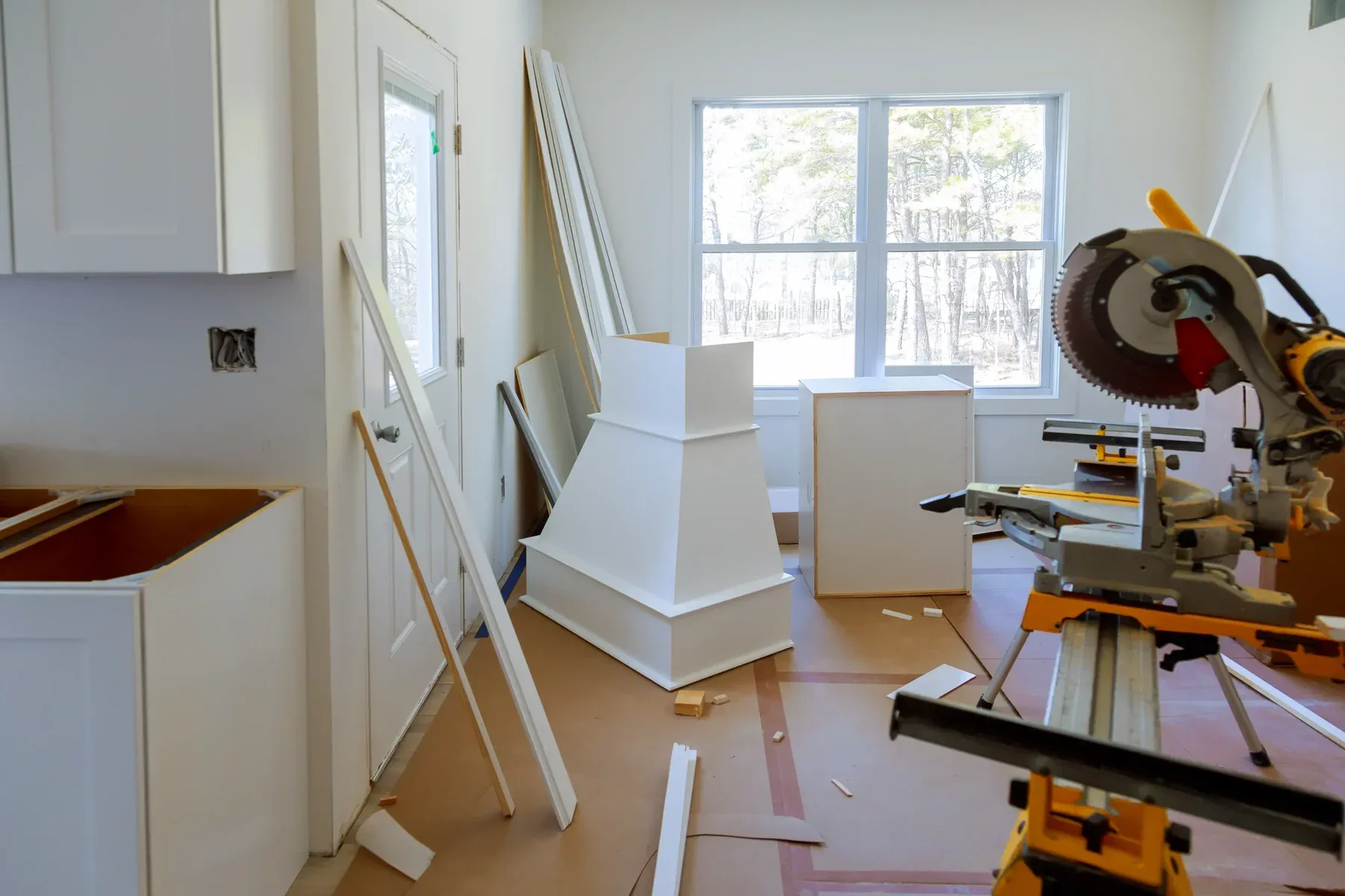 Kitchen under construction with white cabinets, a miter saw, and a window.