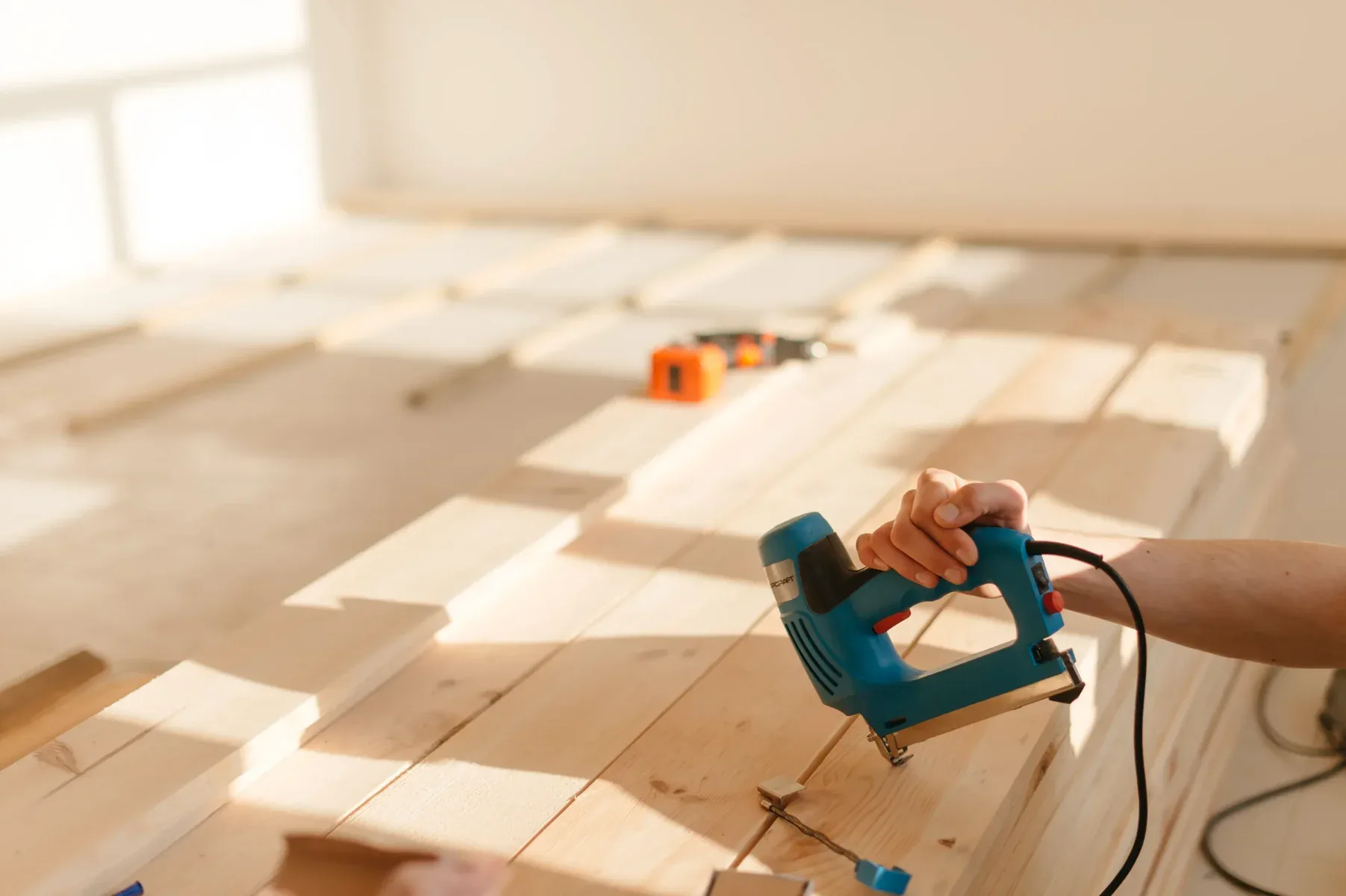 Hand using a blue electric staple gun on light wooden planks.