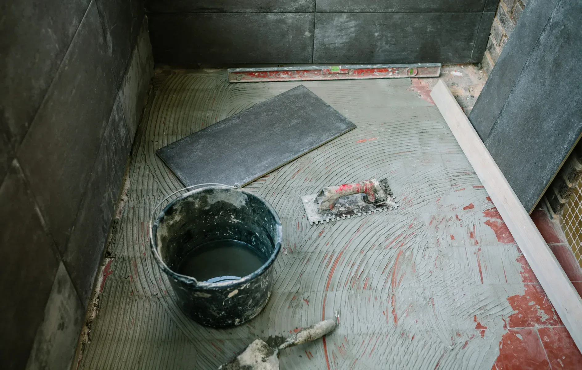Tiling in progress with a bucket, trowel, and dark grey tiles.