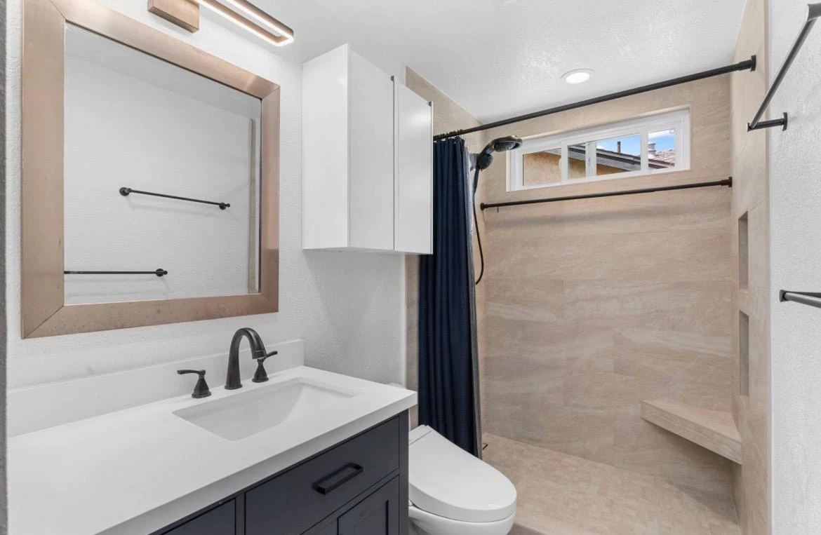 Modern bathroom with dark vanity, white sink, large mirror, and tiled shower.