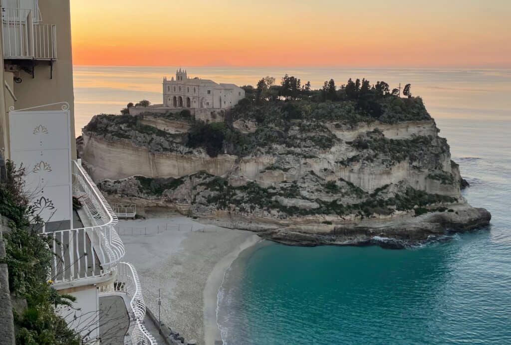 Historic church of Santa Maria della Isola in Tropea on rocky outcrop with white sand beach and colourful beach umbrellas.