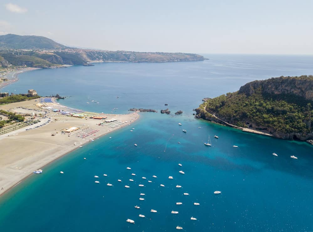 Aerial view of sandy beach left and an island on the right with the blue sea between and the distant mountainous coastline in the distance on a sunny day