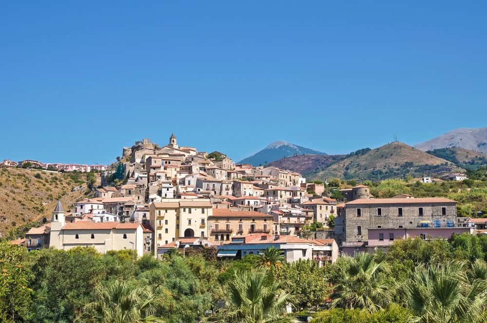 A historic village with pastel coloured houses perched on a mountain top with the buildings stepping down the slope on a sunny day.