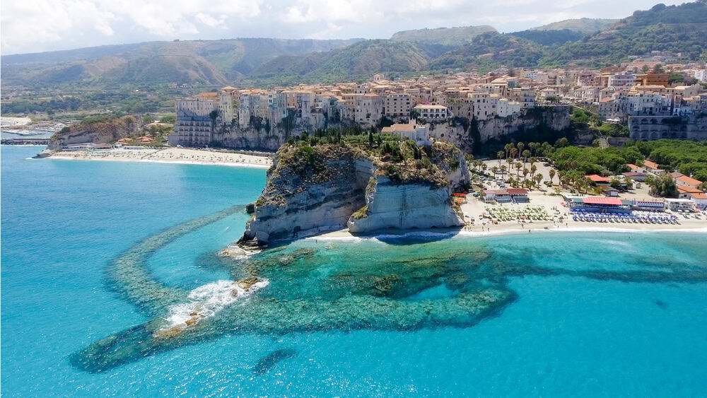 Aerial view of white sand beaches in Tropea with bathers and people on the beaches and the medieval buildings perched on the sheer cliffs above. there is a turquoise blue sea on a beautiful sunny day. and the green montains in the backround above.