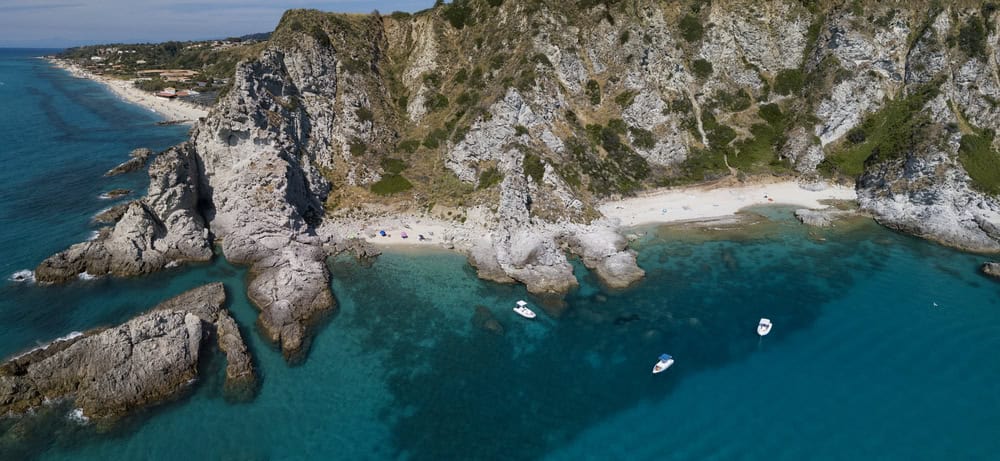 isloated white sand beaches and rocky outcrops with rocky cliffs of Capo vaticano above on a sunny day. Clear blue sea and several anchored boats.