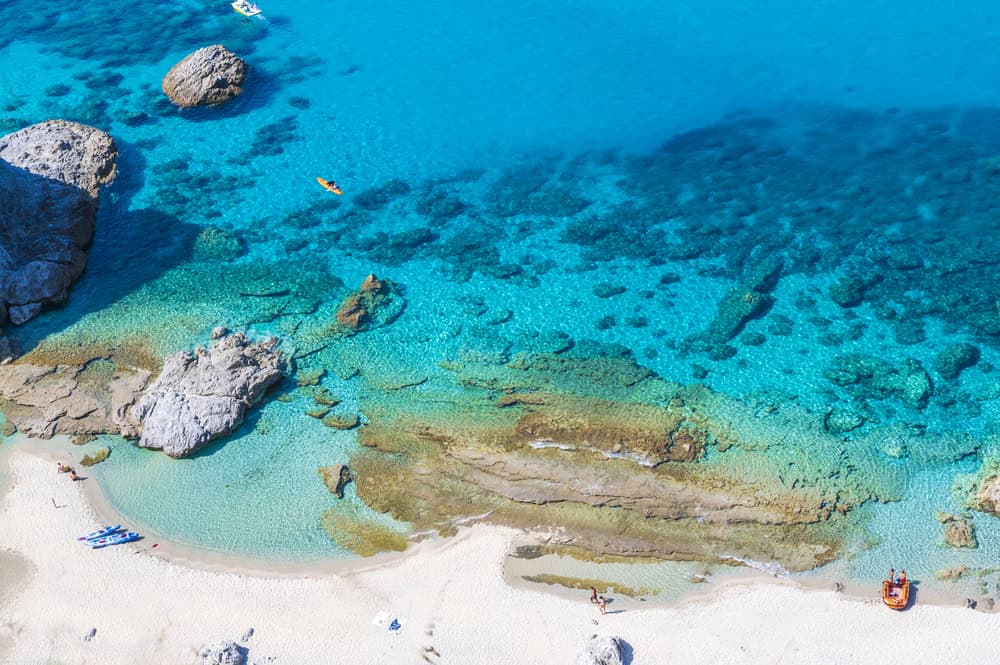 Aerial view of praia di fuoco beach, from the cliffs above. White sand beach with clear turquoise blue sea and reefs visible below. Two kayaks and one small inflatable boat on the shore with two people walking on the beach on a sunny day.
