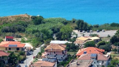 road to a parking area with several houses and green bushland with the blue sea in th background